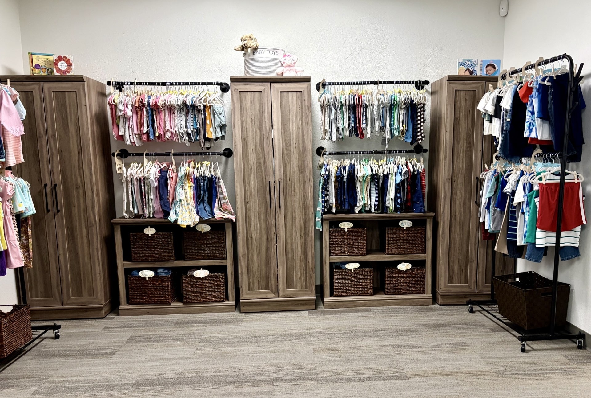 Organized baby and children’s clothing displayed on racks with storage baskets, toys, and books in a family resource center.